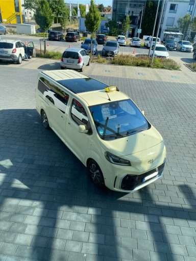 Gelber Minivan mit Taxischild parkt auf einem Parkplatz in städtischer Umgebung.